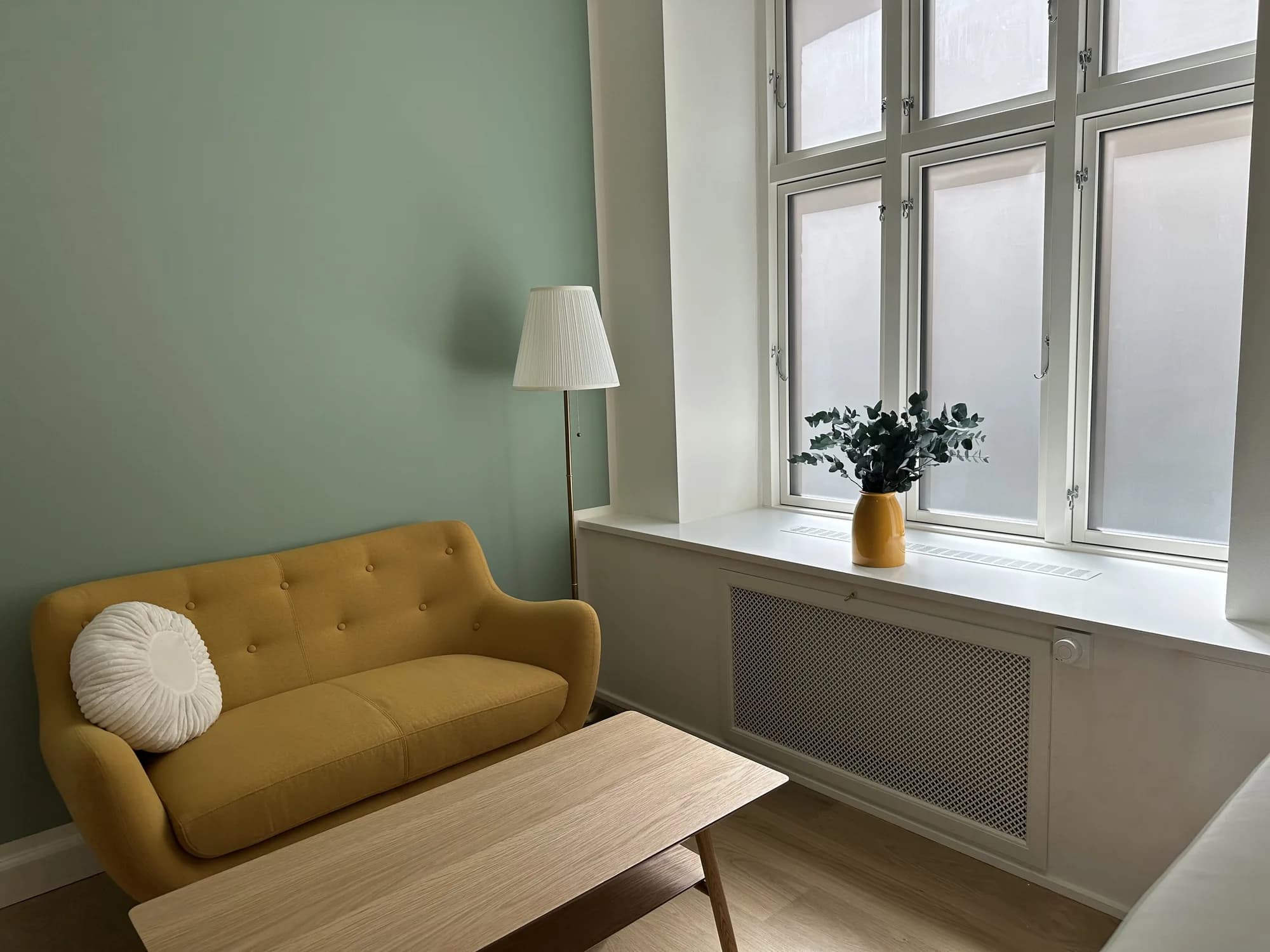 A mustard sofa with a knitted cushion beneath a floor lamp by the frosted window