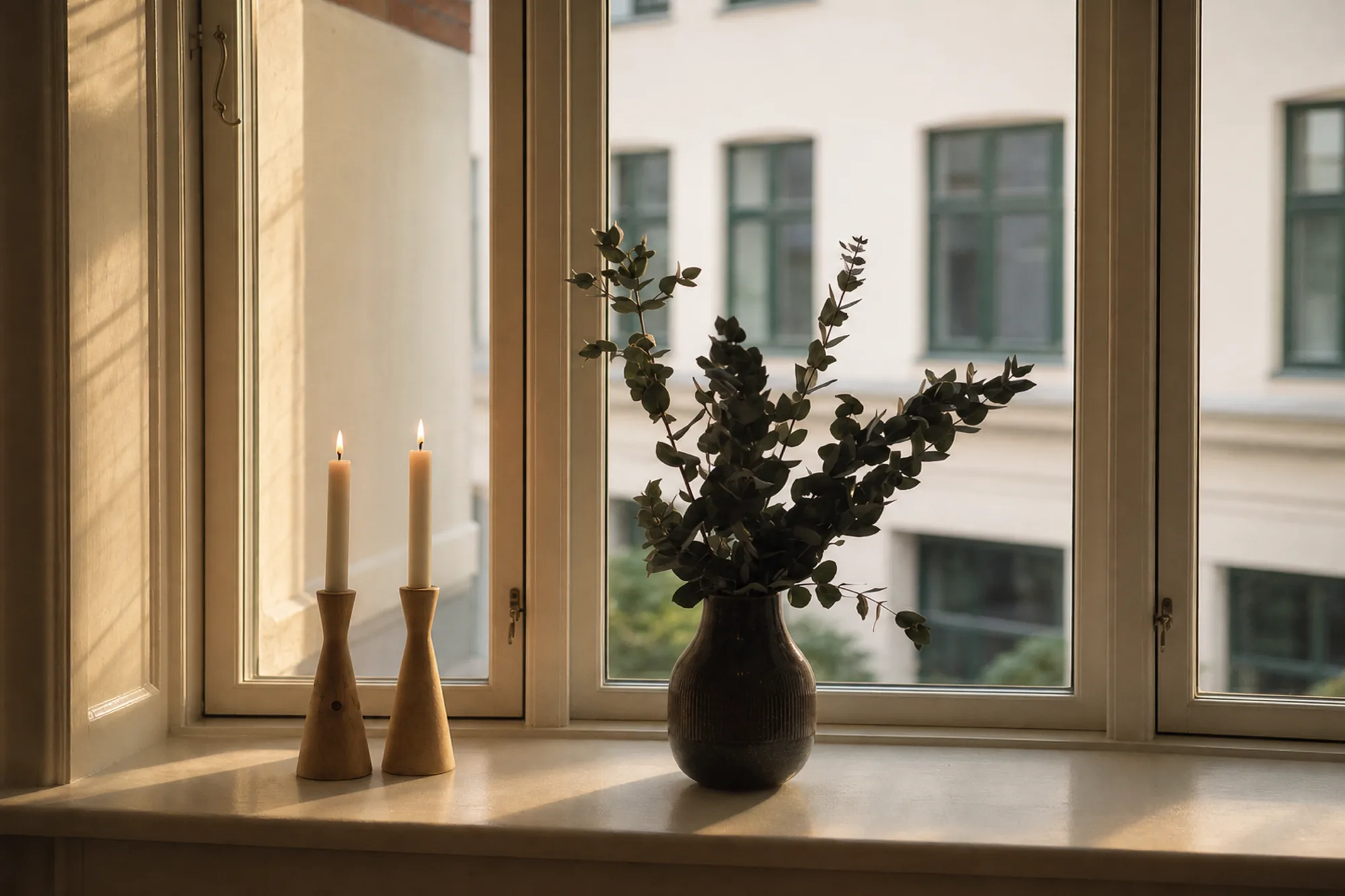 A window sill with a dark vase of branches and two lit candles in golden evening light